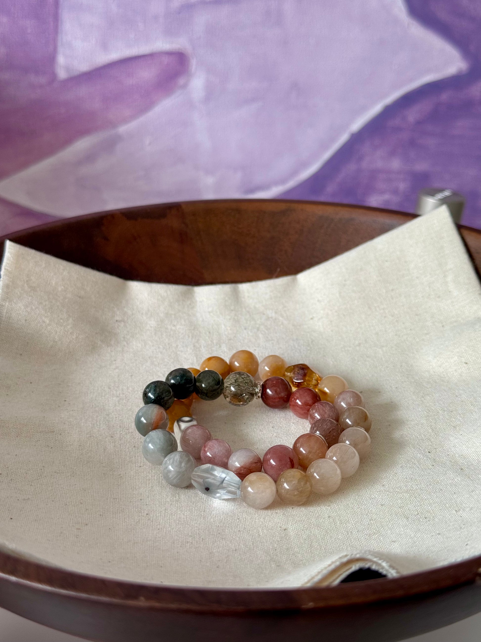 Multicolored beaded rutile quartz bracelet on a wooden tray with a purple fabric background